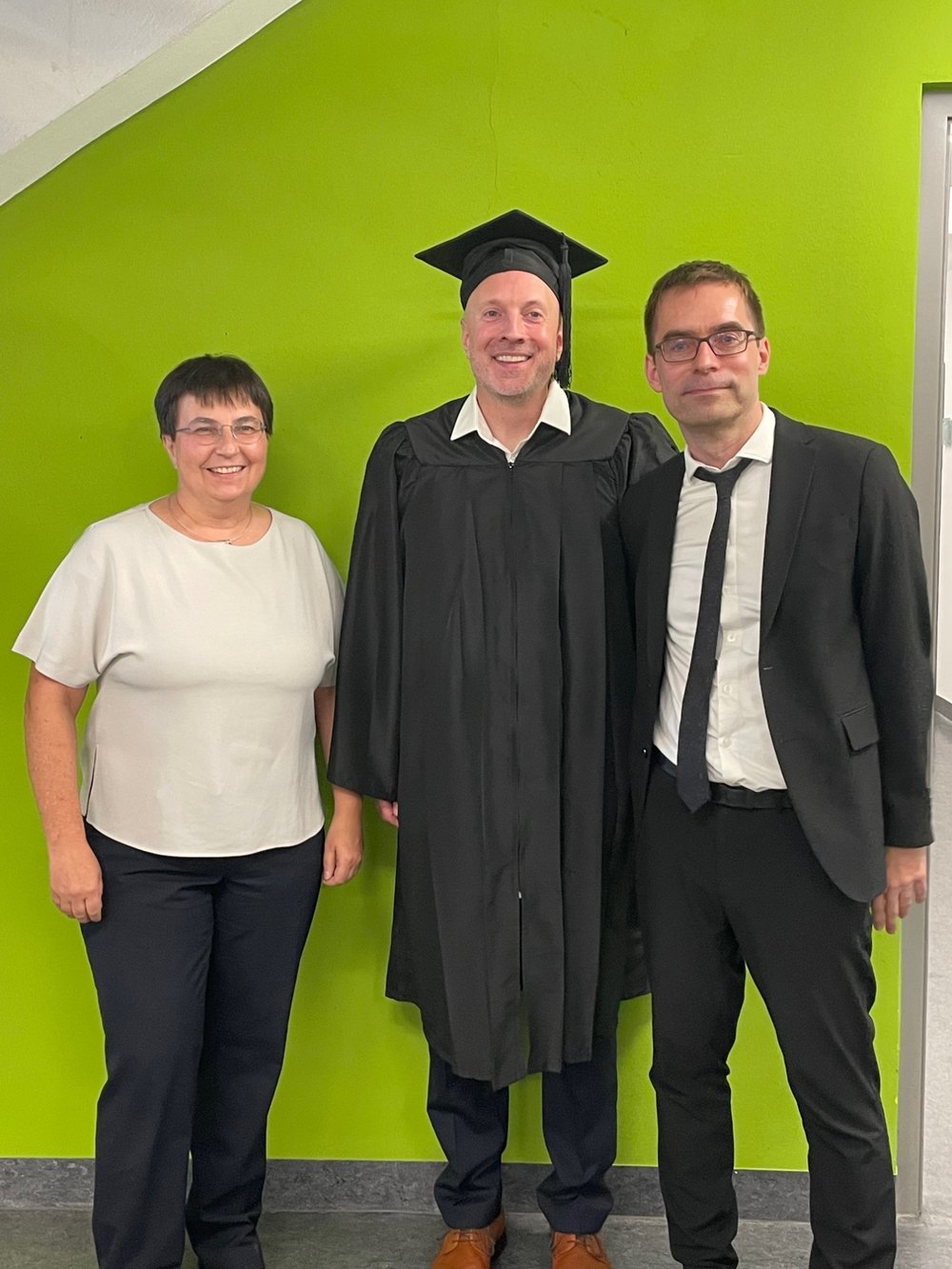 Barbara Dinter, Sebastian Hüttemann in cap and gown, and Roland M. Mueller in front of a green wall