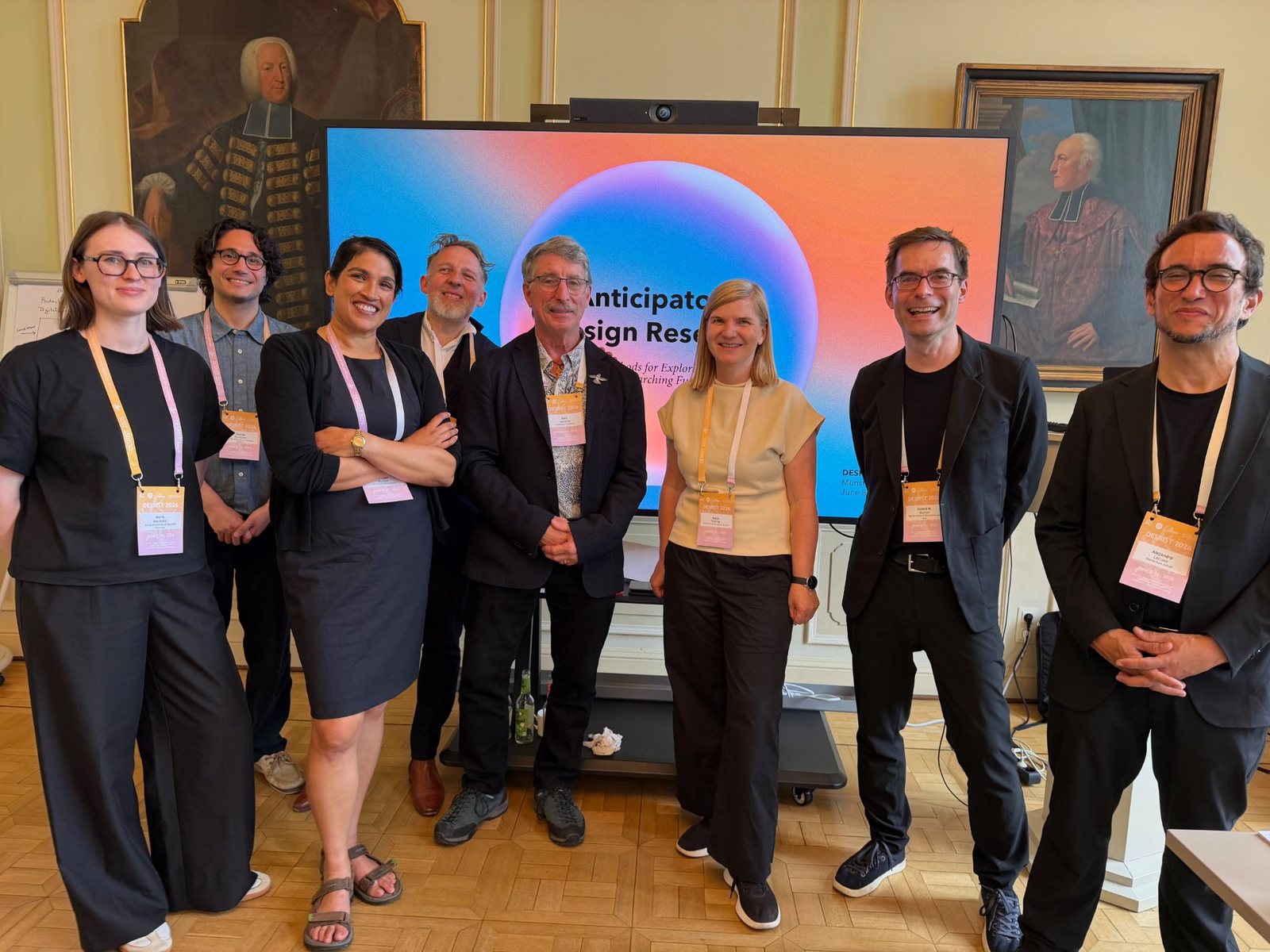 Eight workshop organisers with DESRIST 2026 badges in front of a screen showing the title Anticipatory Design Research, in a historic room with portrait paintings