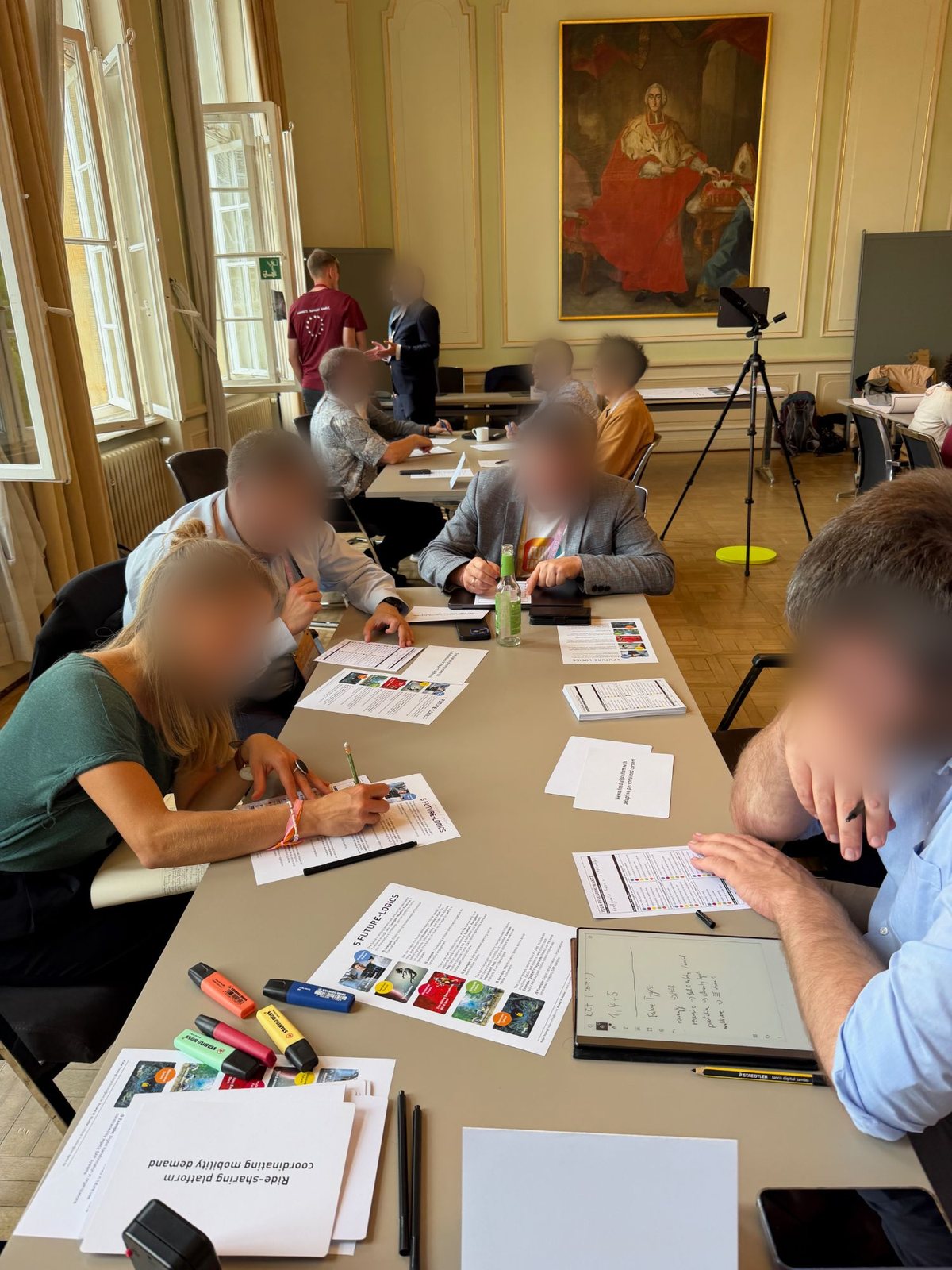 Workshop participants at long tables working with printed Future-Logics sheets, scenario cards such as a ride-sharing platform coordinating mobility demand, and highlighters; faces blurred