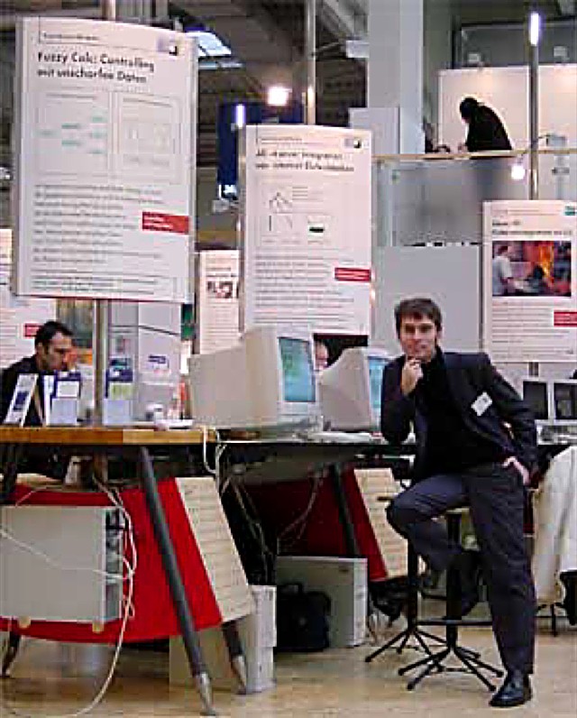 Roland M. Mueller at the FuzzyCalc exhibition stand at CeBIT, next to a poster titled Fuzzy Calc: Controlling mit unscharfen Daten and computer monitors