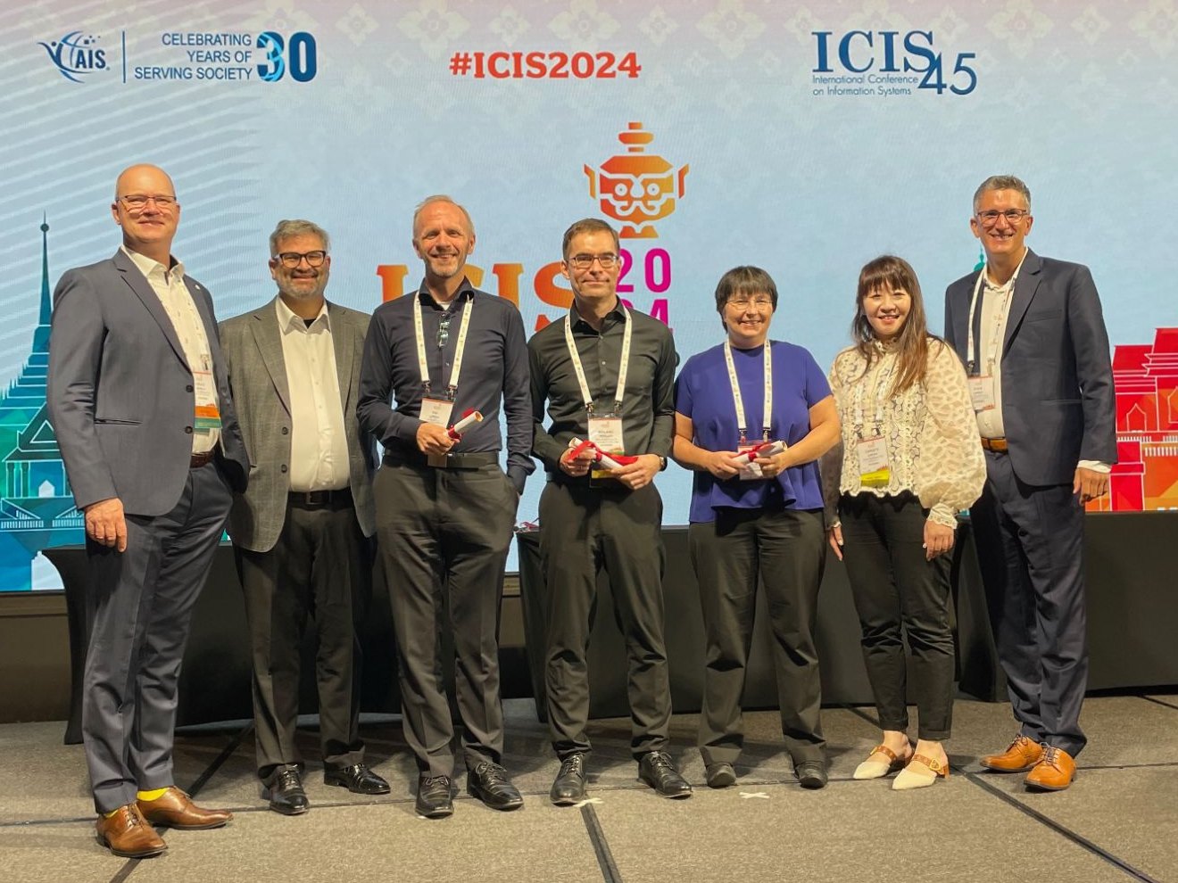 Award ceremony on the ICIS 2024 stage: seven people, three of them holding award certificates, Roland M. Mueller in the centre