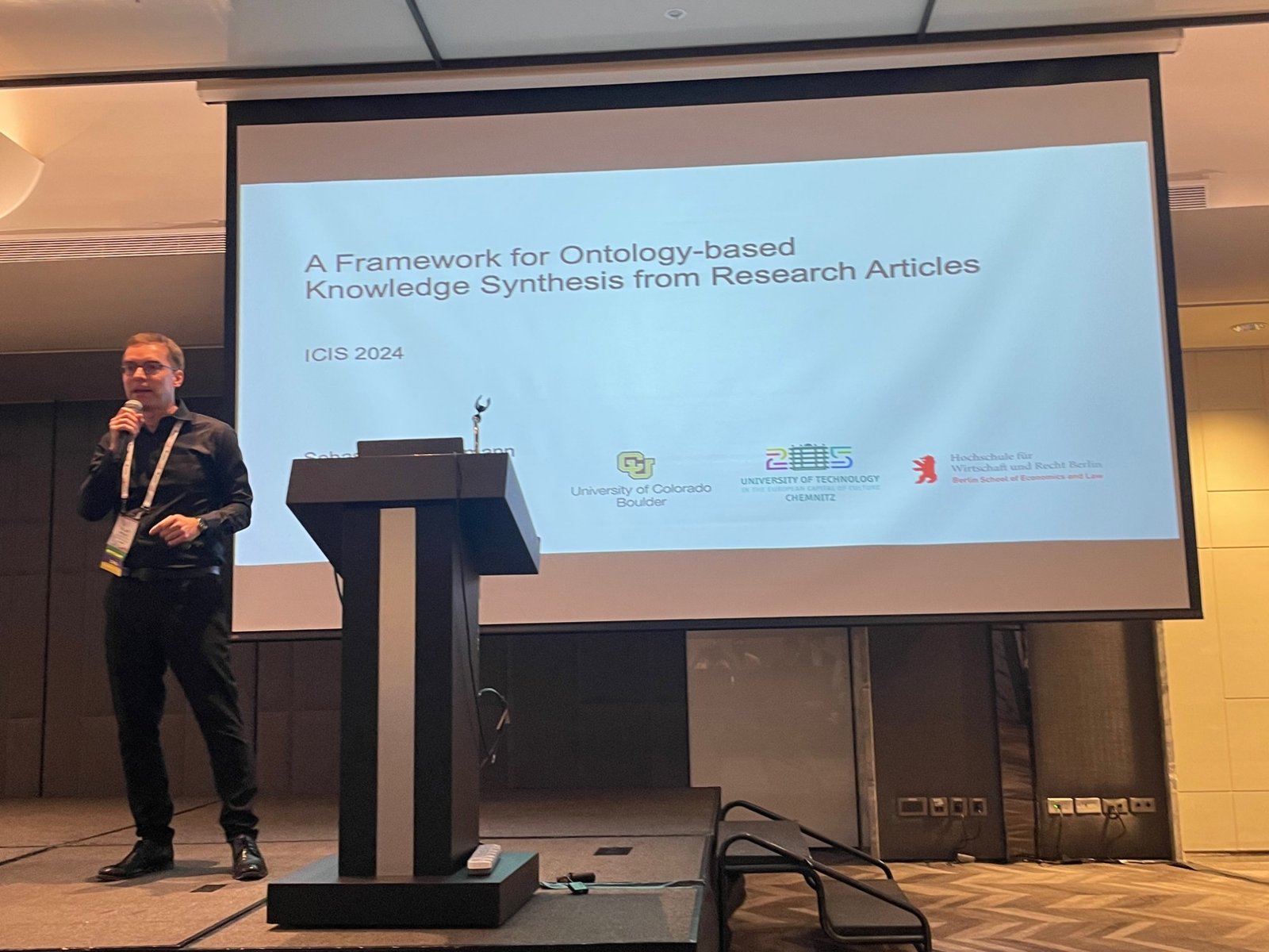 Roland M. Mueller presenting the paper A Framework for Ontology-based Knowledge Synthesis from Research Articles at ICIS 2024, with the title slide and the logos of the University of Colorado Boulder, TU Chemnitz and HWR Berlin