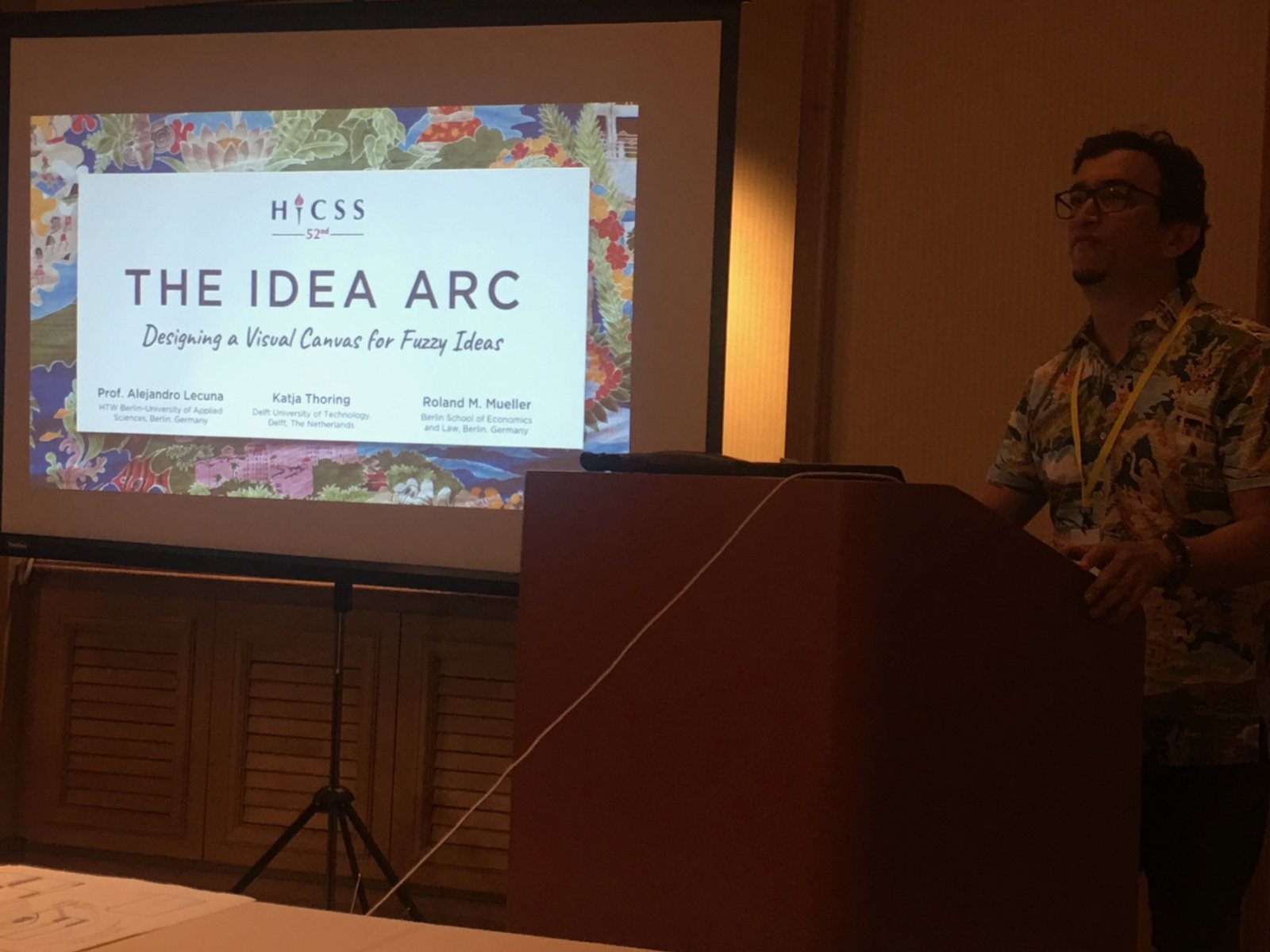 Alejandro Lecuna at a lectern next to a projected title slide: The Idea Arc, Designing a Visual Canvas for Fuzzy Ideas, HICSS 52nd, with the authors Alejandro Lecuna, Katja Thoring and Roland M. Mueller