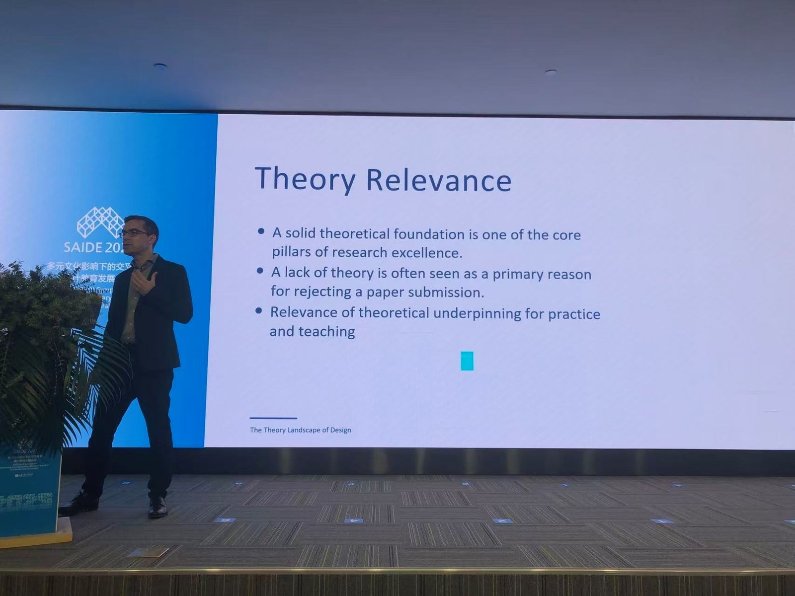 Roland M. Mueller on stage at the SAIDE symposium, presenting a slide titled Theory Relevance from the keynote The Theory Landscape of Design